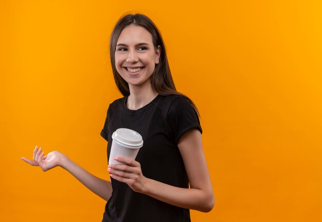 Smiling young caucasian girl wearing black t-shirt looking at side holding cup of coffee on isolated orange background