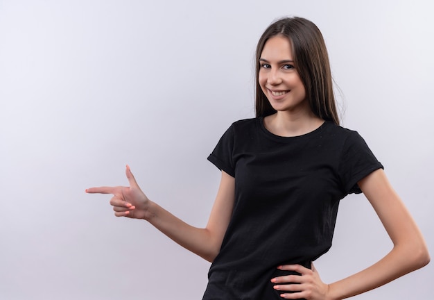 Smiling young caucasian girl wearing black t-shirt points finger to side put her hand on hip on isolated white background