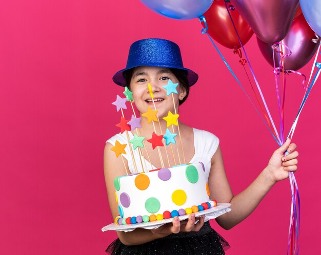 コピースペースでピンクの壁に分離されたバースデーケーキとヘリウム風船を保持している青いパーティーハットを身に着けている若い白人の女の子の笑顔