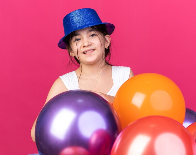 smiling young caucasian girl wearing blue party hat standing with helium balloons  isolated on pink wall with copy space