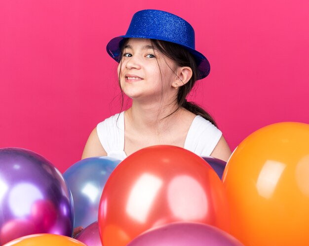 smiling young caucasian girl wearing blue party hat standing with helium balloons isolated on pink wall with copy space