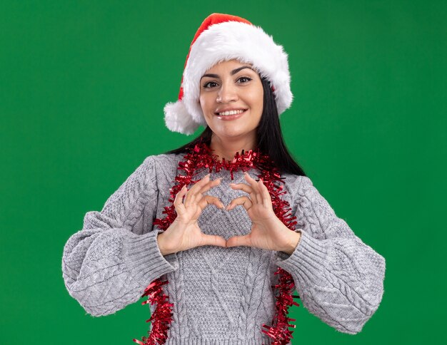 Smiling young caucasian girl wearing christmas hat and tinsel garland around neck looking at camera doing heart sign isolated on green background