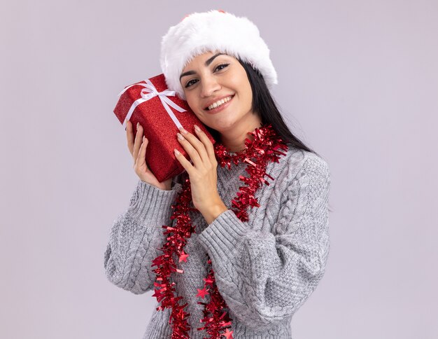 복사 공간 흰색 배경에 고립 된 선물 패키지 감동 머리를 들고 카메라를보고 목에 크리스마스 모자와 반짝이 갈 랜드를 입고 웃는 젊은 백인 여자