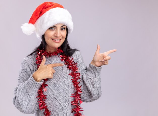 복사 공간 흰색 배경에 고립 된 측면에서 가리키는 카메라를보고 목에 크리스마스 모자와 반짝이 갈 랜드를 입고 웃는 젊은 백인 여자