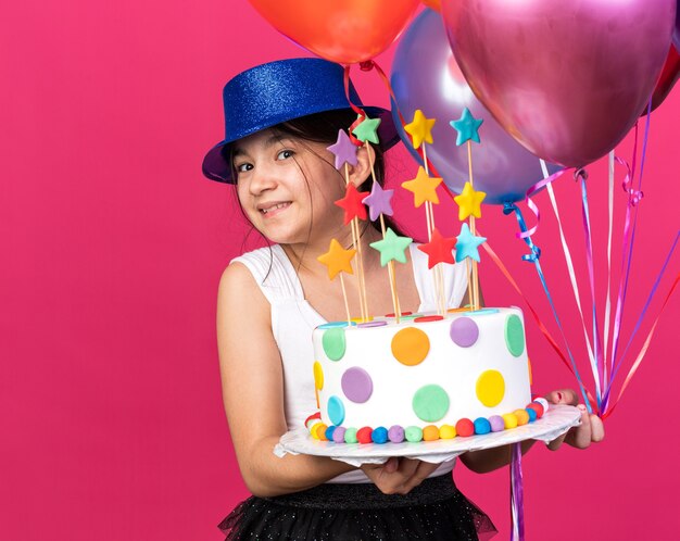 smiling young caucasian girl with blue party hat holding birthday cake and helium balloons isolated on pink wall with copy space