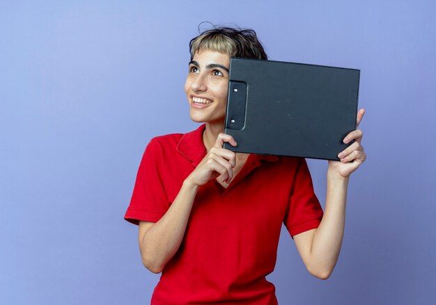 Smiling young caucasian girl with pixie haircut holding clipboard touching face with it and looking up isolated on purple background with copy space