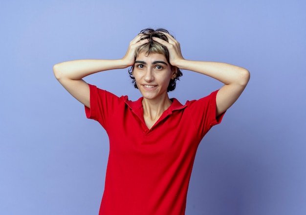 Smiling young caucasian girl with pixie haircut holding head isolated on purple background