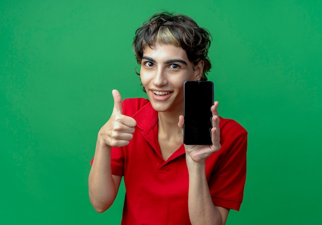 Улыбающаяся молодая кавказская девушка со стрижкой пикси держит мобильный телефон и показывает большой палец вверх на зеленом фоне с копией пространства