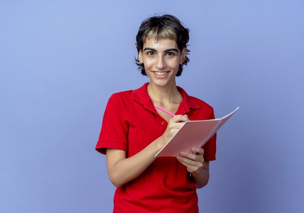 Smiling young caucasian girl with pixie haircut holding pen and note pad writing something isolated on purple background with copy space