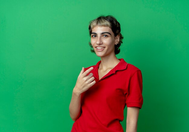 Smiling young caucasian girl with pixie haircut keeping hand in air isolated on green background with copy space