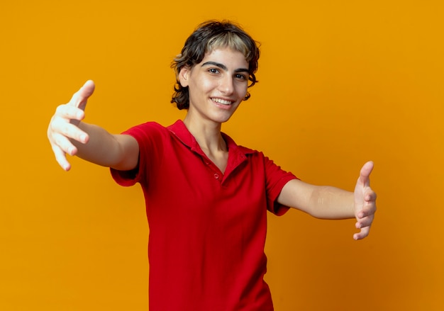 Smiling young caucasian girl with pixie haircut pretend holding something isolated on orange background