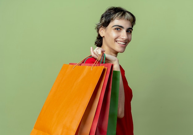 Smiling young caucasian girl with pixie haircut standing in profile view holding shopping bags on shoulder isolated on olive green background with copy space