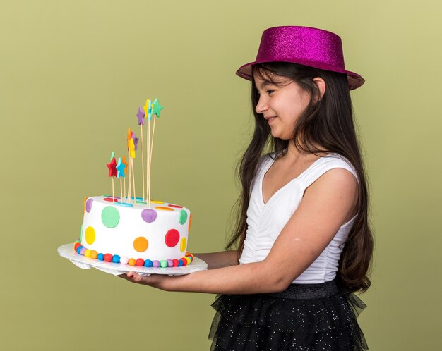コピースペースとオリーブグリーンの壁に隔離された横に立っているバースデーケーキを保持し、見て紫色のパーティハットと笑顔の若い白人の女の子