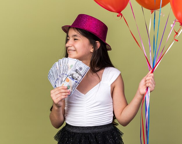 улыбающаяся молодая кавказская девушка в фиолетовой партийной шляпе держит деньги и гелиевые шары, глядя в сторону, изолированную на оливково-зеленой стене с копией пространства