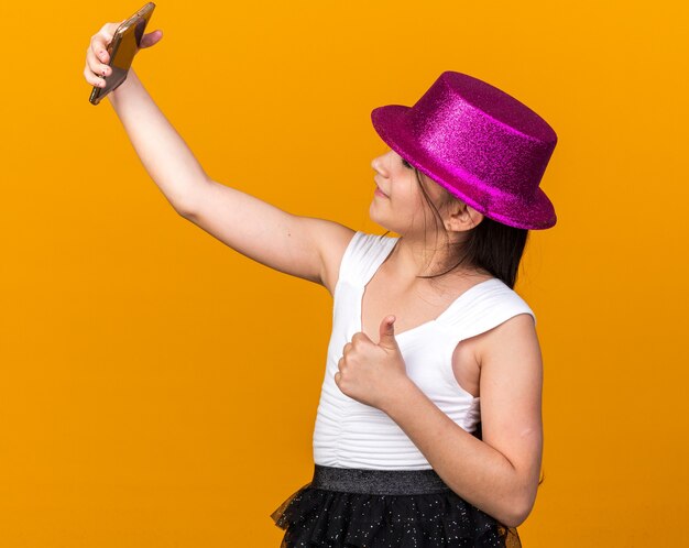 smiling young caucasian girl with purple party hat thumbs up taking selfie on phone isolated on orange wall with copy space