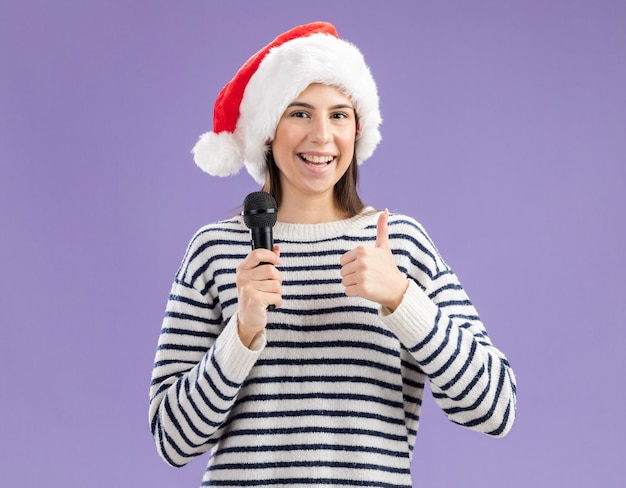 Smiling young caucasian girl with santa hat holds mic and thumbs up isolated on purple wall with copy space