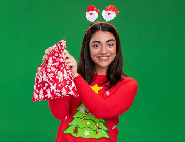 Smiling young caucasian girl with santa headband holding christmas gift bag isolated on green background with copy space