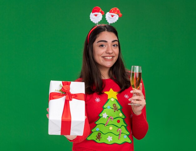 Smiling young caucasian girl with santa headband holds glass of champagne and christmas gift box isolated on green background with copy space