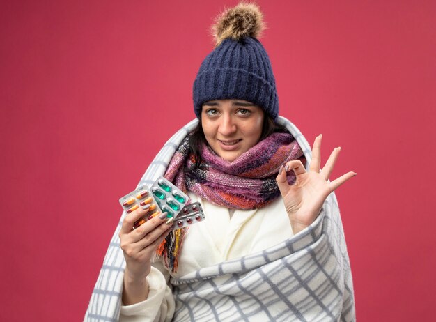 Smiling young caucasian ill girl wearing robe winter hat and scarf wrapped in plaid holding packs of capsules doing ok sign isolated on crimson wall