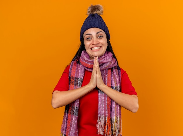 Улыбающаяся молодая кавказская больная девушка в зимней шапке и шарфе держит руки вместе изолированными на оранжевой стене с копией пространства