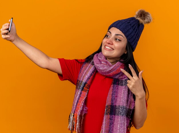 Smiling young caucasian ill girl wearing winter hat and scarf taking selfie doing peace sign isolated on orange wall