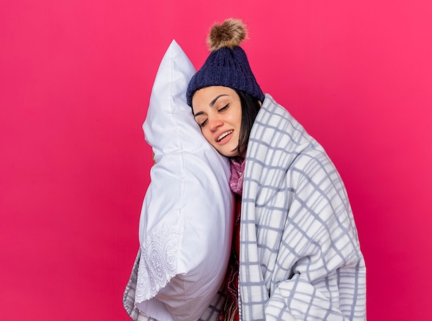 Smiling young caucasian ill girl wearing winter hat and scarf wrapped in plaid hugging pillow putting head on it with closed eyes isolated on crimson wall with copy space