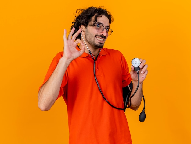 Улыбающийся молодой кавказский больной человек в очках и стетоскопе держит сфигмоманометр, делает хорошо, знак изолирован на оранжевой стене