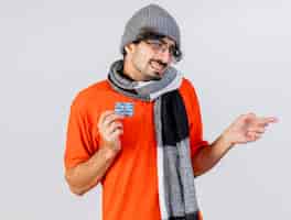 Free photo smiling young caucasian ill man wearing glasses winter hat and scarf holding pack of medical capsules  pointing at side isolated on white wall