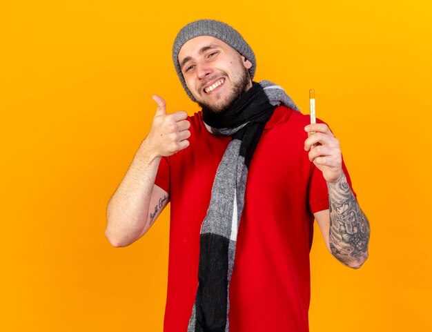 Smiling young caucasian ill man wearing winter hat and scarf thumbs up and holds thermometer