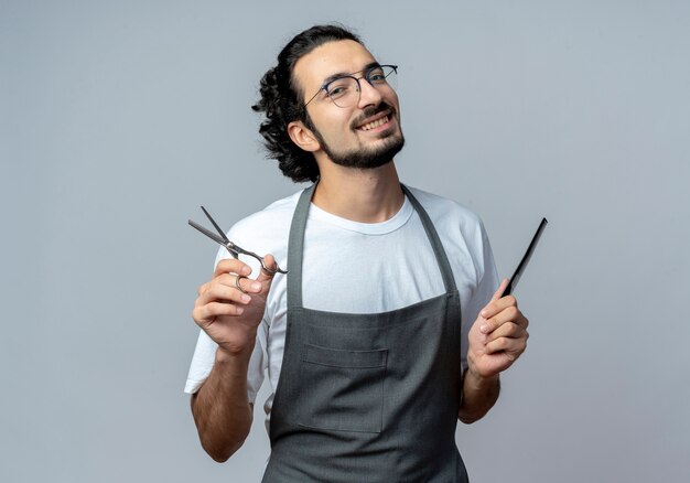 Smiling young caucasian male barber wearing glasses and wavy hair band in uniform holding scissors and comb isolated on white background with copy space