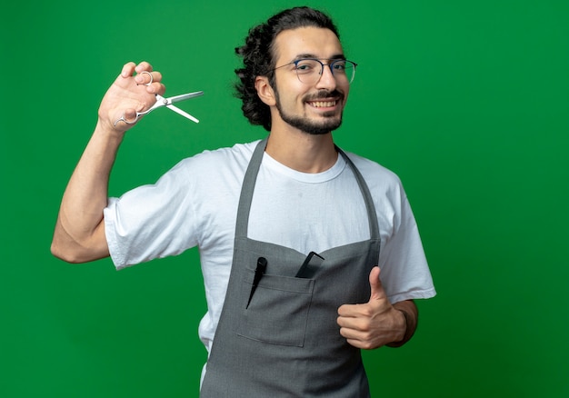 Smiling young caucasian male barber wearing glasses and wavy hair band in uniform holding scissors and showing thumb up isolated on green background with copy space