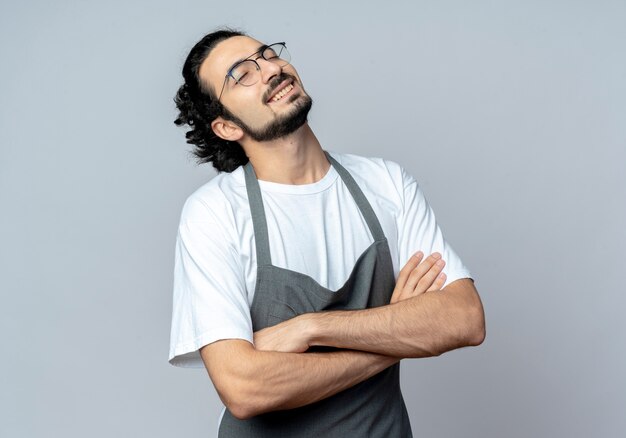 Smiling young caucasian male barber wearing glasses and wavy hair band in uniform standing with closed posture with closed eyes isolated on white background with copy space