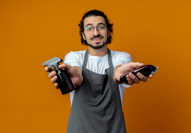 Smiling young caucasian male barber wearing glasses and wavy hair band in uniform stretching out spray bottle and hair clippers