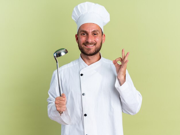 요리사 유니폼을 입은 웃고 있는 백인 남성 요리사와 모자를 들고 올리브 녹색 벽에 격리된 확인 표시를 하는 카메라를 바라보는 모자