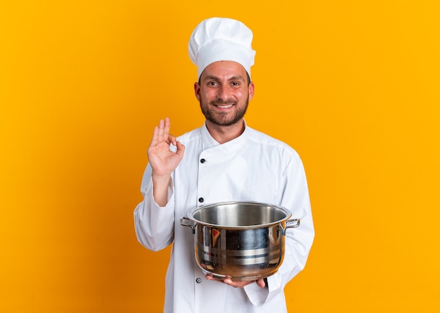요리사 유니폼을 입은 웃고 있는 백인 남성 요리사와 확인 표시를 하는 냄비를 들고 있는 모자