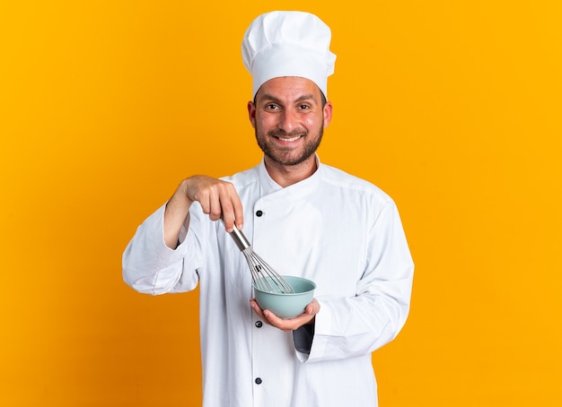 Smiling young caucasian male cook in chef uniform and cap looking at camera whisking eggs with whisk in bowl isolated on orange wall