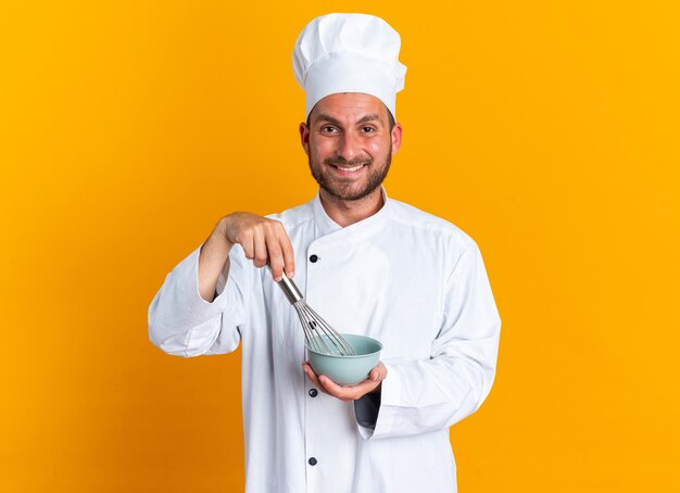 Улыбающийся молодой кавказский мужчина-повар в униформе шеф-повара и кепке смотрит в камеру, взбивая яйца венчиком в миске, изолированной на оранжевой стене
