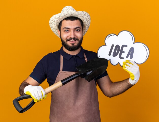 smiling young caucasian male gardener wearing gardening hat and gloves holding and pointing at idea bubble with spade isolated on orange wall with copy space