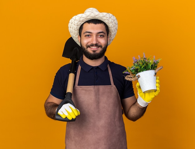 smiling young caucasian male gardener wearing gardening hat and gloves holding spade on shoulder and flowerpot isolated on orange wall with copy space