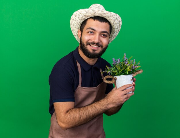 Sorridente giovane maschio caucasico giardiniere indossando giardinaggio hat holding vaso di fiori isolato sulla parete verde con copia space