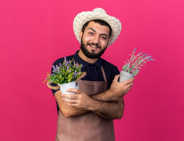 smiling young caucasian male gardener wearing gardening hat holding flowerpots crossing arms isolated on pink wall with copy space