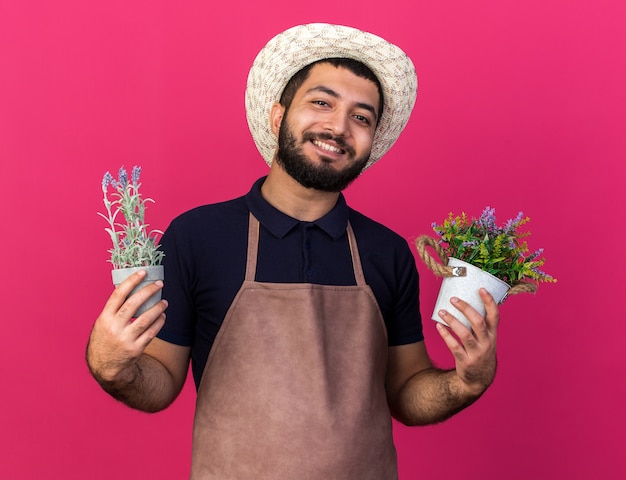 Foto gratuita sorridente giovane maschio caucasico giardiniere che indossa cappello da giardinaggio tenendo vasi di fiori isolati sulla parete rosa con spazio di copia