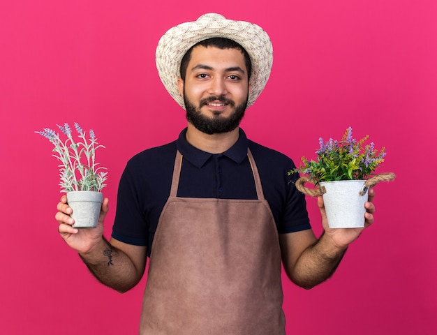smiling young caucasian male gardener wearing gardening hat holding flowerpots isolated on pink wall with copy space