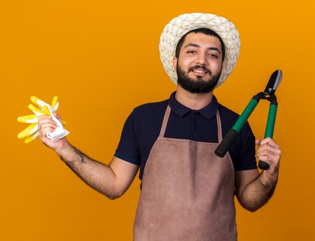 smiling young caucasian male gardener wearing gardening hat holding gardening scissors and gloves isolated on orange wall with copy space