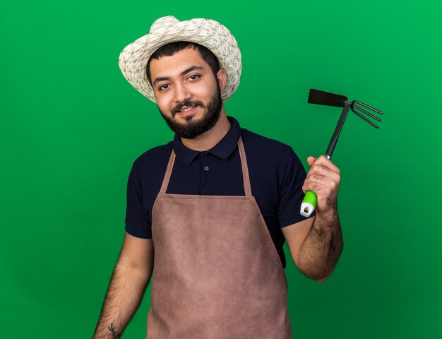 smiling young caucasian male gardener wearing gardening hat holding hoe rake isolated on green wall with copy space