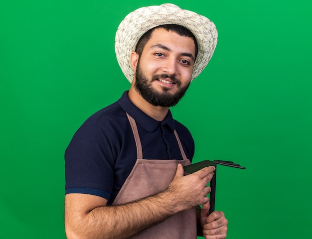 smiling young caucasian male gardener wearing gardening hat holding hoe rake isolated on green wall with copy space