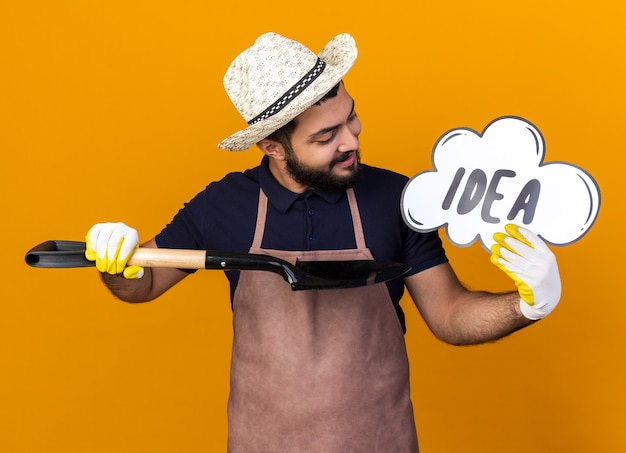 Foto gratuita sorridente giovane maschio caucasico giardiniere che indossa cappello da giardinaggio guardando e puntando a idea bolla con vanga isolata sulla parete arancione con spazio di copia