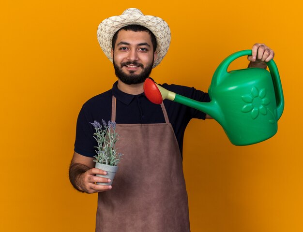 smiling young caucasian male gardener wearing gardening hat pretending to water flowers in flowerpot with watering can isolated on orange wall with copy space