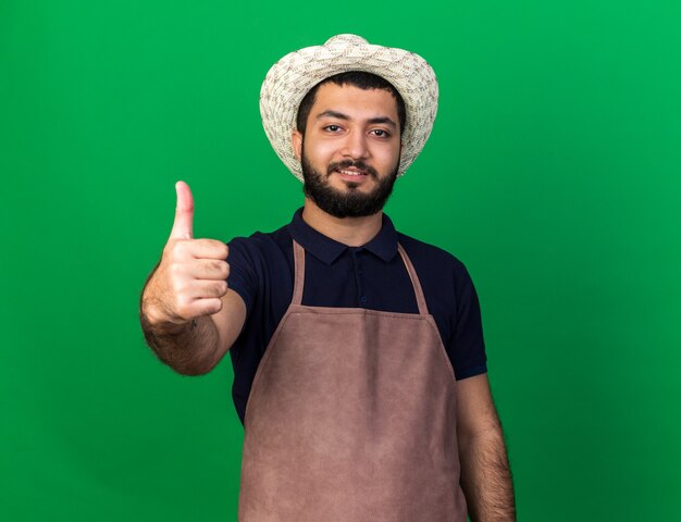 smiling young caucasian male gardener wearing gardening hat thumbing up isolated on green wall with copy space