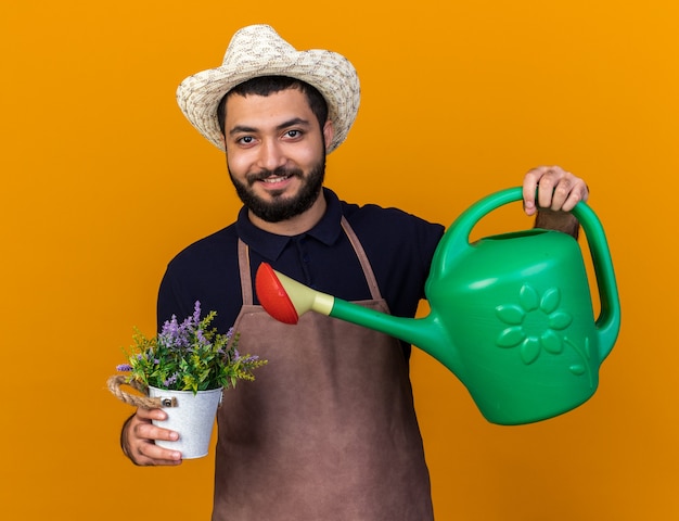 ガーデニング帽子をかぶって笑顔の若い白人男性の庭師は、コピースペースでオレンジ色の壁に分離することができます散水と植木鉢の花に水をまきます